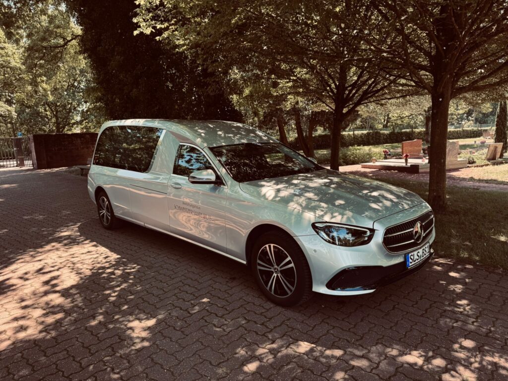 Silberner Mercedes E-Klasse Bestattungswagen des Bestattungshauses Schweitzer unter Bäumen auf einem Friedhof bei Saarlouis