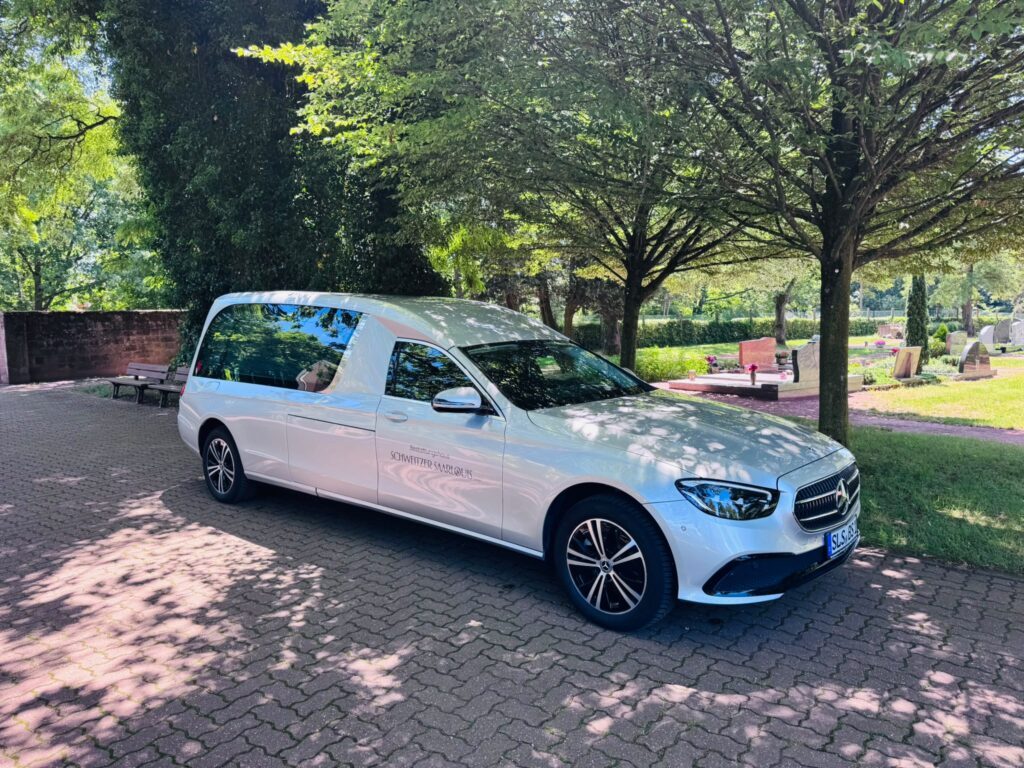 Weißer Mercedes E-Klasse Bestattungswagen des Bestattungshauses Schweitzer Saarlouis auf einem Friedhofsvorplatz