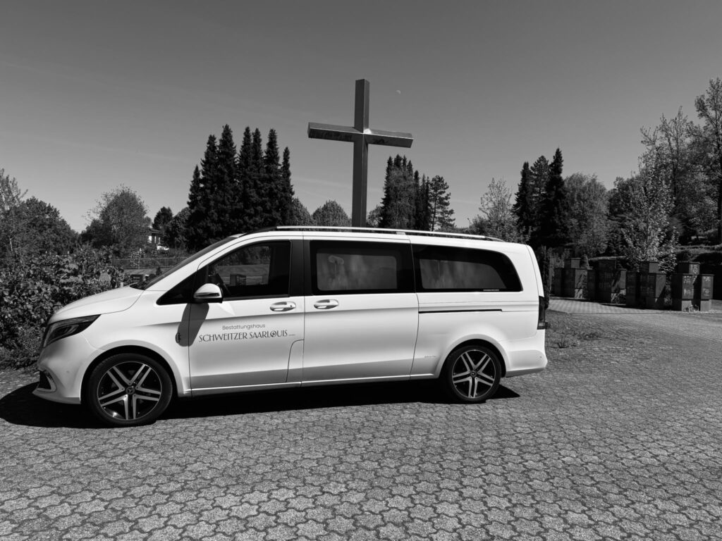 Mercedes V-Klasse Bestattungswagen des Bestattungshauses Schweitzer vor einem Friedhofskreuz, Schwarzweiß-Aufnahme